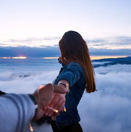 A photo of a man’s hand extended to hold a woman’s hand with an overlooking view of mountains and clouds in the background