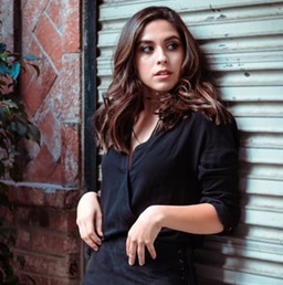 A photo of a Latina in a black top and black skirt, leaning against a wall